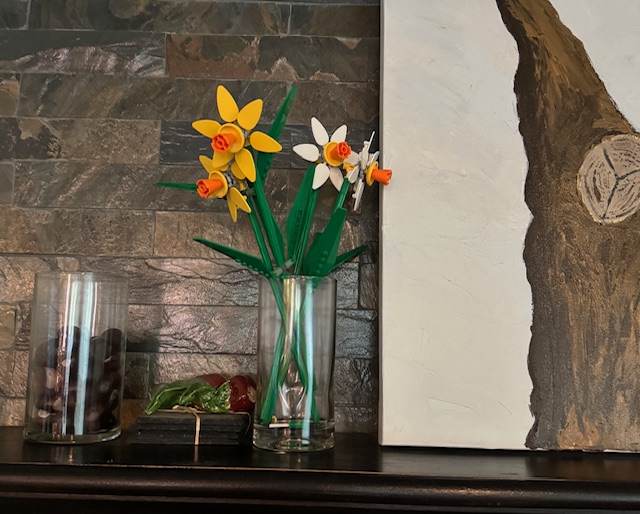 LEGO daffodils, placed in a glass jar on top of a mantel, next to a painting of a tree.