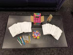 A tray is set with fanned out burp cloths. In the center, is a bowl filled with various foam stamps with shapes of flowers and leaves on them. Markers are spread out next to the bowl of stamps, and there's a box of paints and paint brushes in a jar.