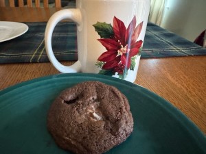 One chocolate cookie with a bit of marshmallow showing through. It's on a plate with a mug in the background. The mug has a poinsettia painted on it.