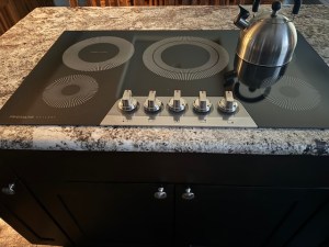 Obsidian-colored glass cooktop with silver knobs. The burners are outlined in white. The cooktop is set on a kitchen island with cabinets beneath, and there's a silver-colored teapot on one of the burners.