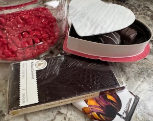 This photograph shows a countertop with an assortment of candies, arranged artistically. There's an opened box of chocolates, and the box is in a heart shape. There is one solid chocolate bar that is sea salt flavored, and there is another chocolate bar under it. Most of the label is covered, except for a beautiful red flower against a white background.