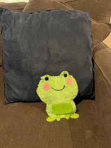 This photo pictures a stuffed frog with some seams showing on the front. It's a kind of felt frog with a fluffy front. It has pink, rosy cheeks, black eyes, and a piece of black thread for the smile. It is propped up against other pillows on a couch.