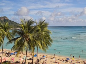 This photo shows a balcony view of Waikiki Beach on a bright sunny day. Palm trees are in the foreground, and tourists in colorful bathing suits dot the beach and the turquoise blue water in the background.
