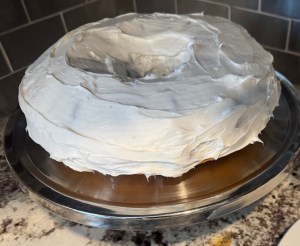 This photo displays a doughnut-shaped vanilla Bundt cake with white frosting. The cake is placed on a silver-colored cake stand on a marble countertop.
