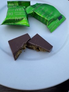 This photo displays a square of "Dubai" dark chocolate, broken open on a white plate. The pistachio filling is exposed. Behind this square are two more green foil-wrapped chocolates.