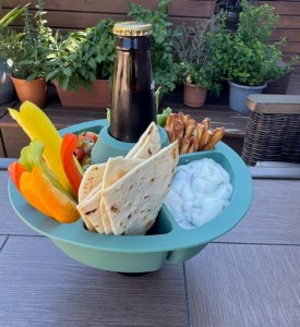 This photo shows a green, silicone chip and dip tray with divided compartments, and there's an opening in the middle to hold a bottled beverage. In this photo, the bottled beverage is a cherry soda, and the dip containers hold colorful pepper strips, flour tortilla wedges (baked), a plain yogurt vegetable dip, pretzel twists, and salsa.
