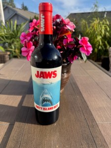 This photo shows a bottle of red wine on a deck in front of a plant, outside in a backyard/neighborhood. The red wine label reads: Jaws: Amity Island Red Blend.