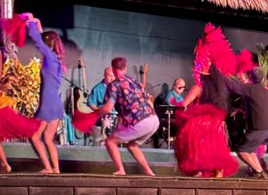 This photo shows more of Alex dancing the hula on stage with other hula dancers in red feathered costumes.