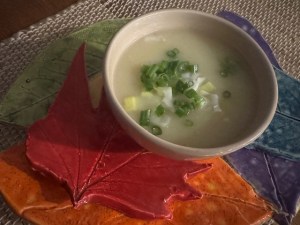 Beige bowl of soup, placed on colorful ceramic leaf shapes. The soup is a potato and leek soup with chives and extra leeks as a garnish.