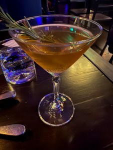 This photo shows the Curtain Call drink at the Kro Bar in Bothell, Washington. It is in a dimly lit, atmospheric room, on top of a dark wood-grain table. The drink glass is martini shaped and the liquid inside is an amber color. There's a sprig of rosemary balanced on the rim.
