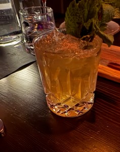 This photo shows a cocktail that's amber-colored in a "rocks glass" with sprigs of mint maybe that are decoratively placed on top.