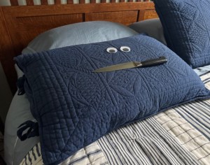 This photo shows a giant blue pillow with big googly eyes and a chef's knife for a mouth. It's placed on a bed with a blue-and-white-striped duvet.