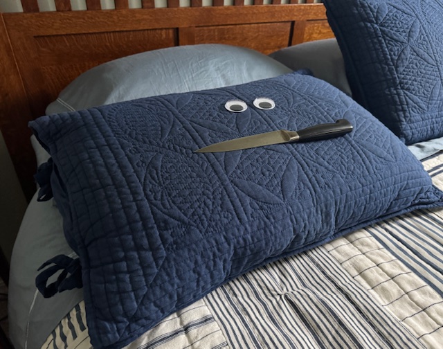 This photo shows a giant blue pillow with big googly eyes and a chef's knife for a mouth. It's placed on a bed with a blue-and-white-striped duvet.