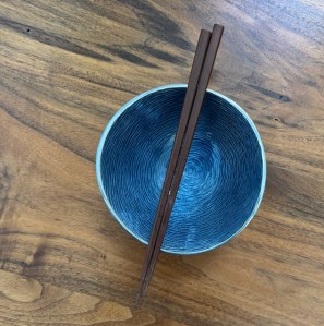 This photo shows a fancy blue bowl with chopsticks lined over the top. The bowl is placed on a wooden table, and the view is from looking down into the bowl/chopsticks.