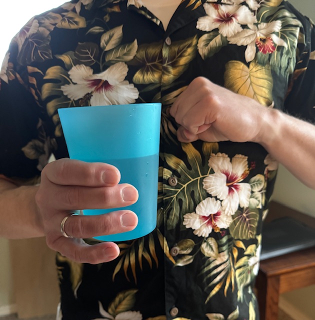 This is a photo of a man in a Hawaiian shirt holding a blue plastic cup of water and making a fist over his chest, like he might be hiccuping.