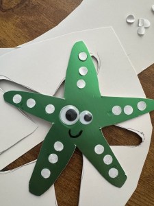 Photo of a shiny green paper sea star with two googly eyes (one is bigger than the other). It is smiling, and it has three little paper dots on each ray/arm. It is set against the paper from which it was cut (the white underside of it). Some extra paper dots are in the background, and it's all set upon a wooden table.