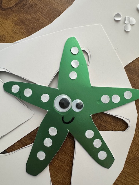 Photo of a shiny green paper sea star with two googly eyes (one is bigger than the other). It is smiling, and it has three little paper dots on each ray/arm. It is set against the paper from which it was cut (the white underside of it). Some extra paper dots are in the background, and it's all set upon a wooden table.