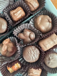 This is a photo of a box of chocolates from the inside. The chocolate pieces are nestled inside individual paper cup wrappers. Some look like chocolate covered nut clusters. Some are round and covered in chocolate. Some are square and covered in chocolate. Both dark and milk chocolate varieties are represented.