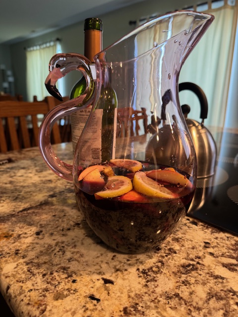 This is a pitcher of a pink tinted flamingo-shaped pitcher that's see-through. It is filled 1/3 of the way with sangria, and you can see slices of citrus and other fruit floating on top. The pitcher is placed on a marble-like countertop with a chair/living room in the background. Directly behind the pitcher, there's a teapot and electric-top stove.