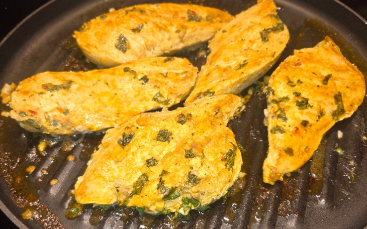 This photo shows boneless, skinless chicken pieces sizzling on a grill pan. They're studded with turmeric and spices and garlic and cilantro.
