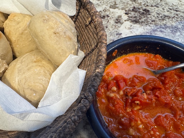 This is a photo of red pepper sauce/dip in a blue Fiesta Ware bowl with a basket of homemade rolls next to it. There's a spoon in the dip, and the items are placed on a marbled countertop.