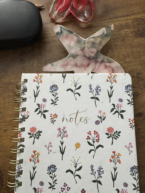 This is a photo of a spiral-bound notebook that says "Notes" in gold ink. There's a pink and green flower motif on the front. Sticking out of the top is a floral starfish bookmark--and it's cut into the shape of a starfish. It is placed on a wooden table, and in the background you can see the edge of an eyeglass case, the swirls of the bottom rays of a glass starfish, and a pencil tip.