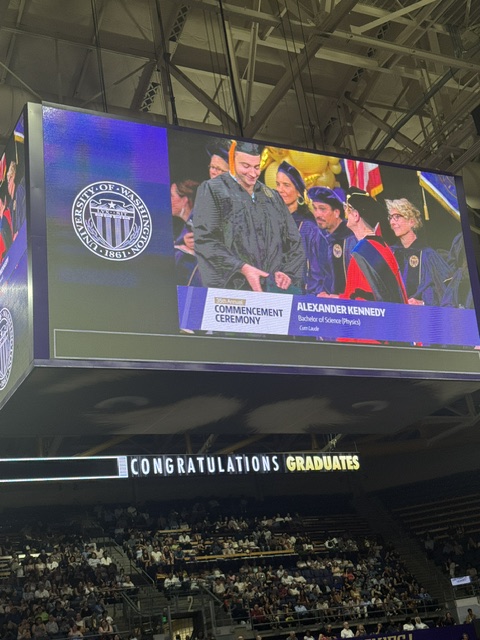 This is an image of Alex walking across the stage to receive his diploma. It's projected onto the JumboTron. He's surrounded by professors in university garb. The banner underneath says Alexander Kennedy, Bachelor of Science in Physics, Cum Laude