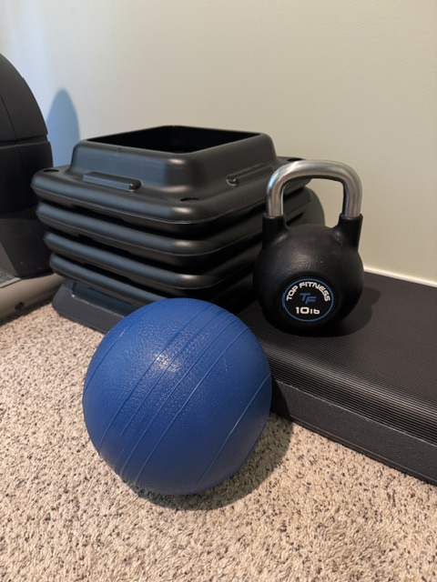 This photo shows a home gym with a step , riser blocks, a blue weighted ball, a kettle weight of ten pounds, and a glimpse of an exercise bike in the background.