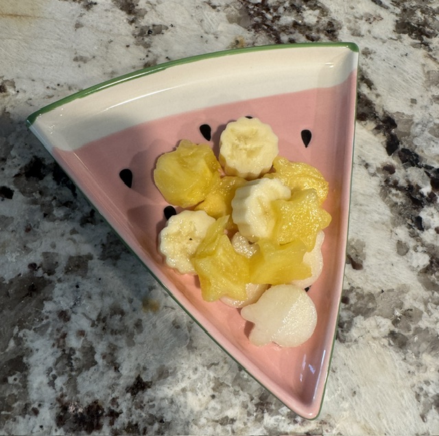 This is a photo of a watermelon slice shaped plate (triangular) with fruit salad placed on it. The fruit is cut into cute shapes. The plate is placed on a marble-like countertop.