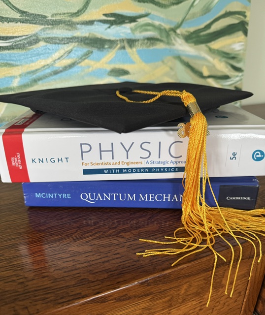 This is a photo of a black graduation cap with a yellow/gold tassel. It is resting atop two textbooks: Physics and Quantum Mechanics. In the background is an abstract painting of green, aqua, and yellow brushstrokes.