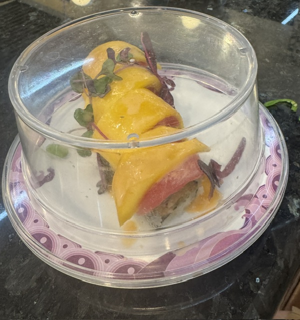 I'm assuming this is a Godzilla roll. It is beautifully presented with maybe fresh mango slices on the top and what looks like tuna and rice inside the roll. It's decorated with fresh sprigs of herbs, and it's placed on a white plate with violet trim. A clear plastic dome covers the dish.