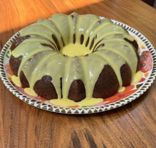 This is a photo of a Bundt cake, placed on a festive plate. Lemon yellow icing drips and gleams down the sides and on the top. The plate is set on a wooden table.