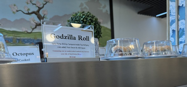 This is a photo of sushi on a conveyor belt at a restaurant. The signs above read Godzilla Roll--and in the back, there's a sign for Octopus.