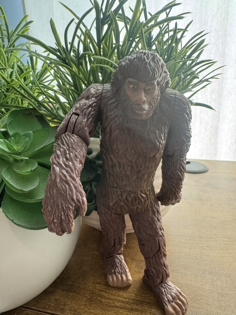Photo of a Bigfoot action figure in front of fake plastic plants on a wooden desk.