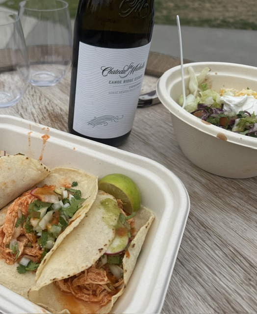 This is a photo of corn tortilla chicken tacos in a styrofoam box. There's a wedge of lime at the top. The tacos are saucy and topped with onion and radish. The box is set upon a wooden wine barrel "table." In the background you can see a picture of Chateau St. Michelle wine, two clear plastic glasses, and a carne asada steak bowl. 