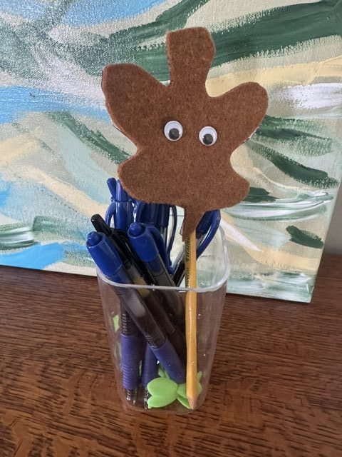 This is a photo of a glass pencil/pen jar that holds blue pens, some tiny 3-D molded rubber leaf-shaped bookmarks, and a pencil with a brown felt leaf-shaped pencil topper with googly eyes.