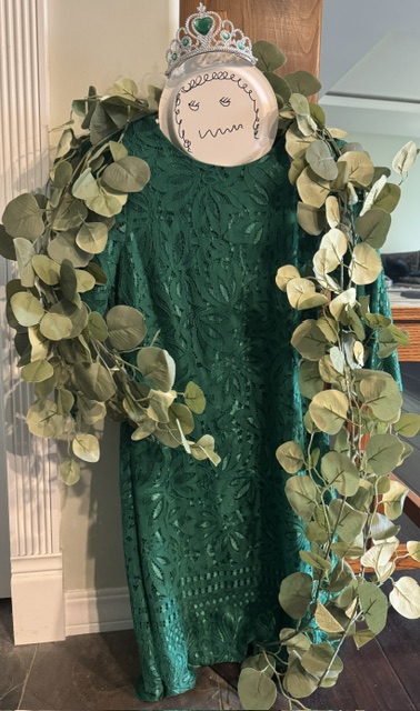 This is a photo of a green dress on a hanger with fake green leaves draped along the shoulders. The dress is hanging from a wooden chair, and there is a textured white wooden trim strip for a wall in the background. Near the neck of the dress, there is a paper plate with a silly face drawn on it and a silver tiara with green gems above the plate/head, balancing on the hanger hook.