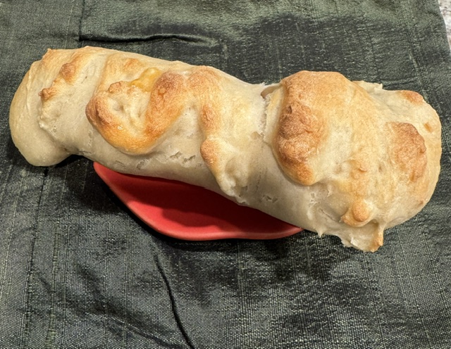 This is a photo of a "mummy" wrapped hot dog on a plate. The strips of dough are hiding the hot dog, and the top is brown. It's resting on a red ceramic plate against a dark green fabric background.